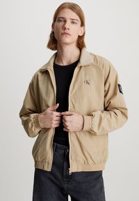 Chaqueta ligera beige con cierre de cremallera frontal, puños acanalados y logotipo bordado. Presenta un ajuste holgado y cintura elástica.
