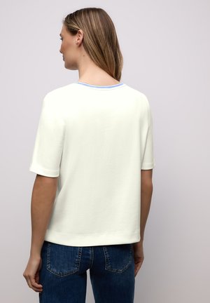 Femme aux longs cheveux portant un t-shirt blanc à manches courtes avec un bord bleu au niveau de l'encolure et un jean bleu, vue de dos sur un fond clair.