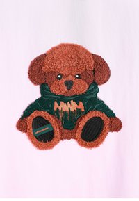 Un oso de peluche marrón con pelaje texturizado, vistiendo un suéter verde con toques naranjas, sentado contra un fondo rosa claro.