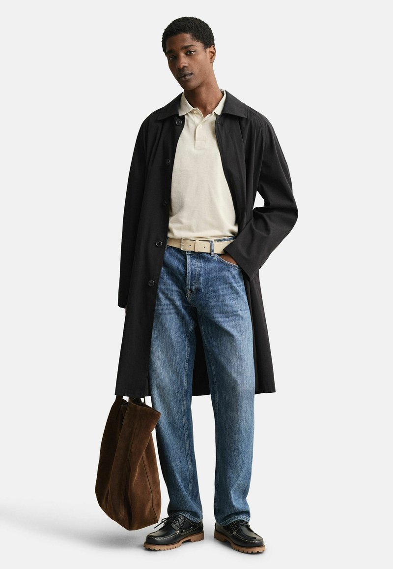 Jonge man met een lange zwarte jas, beige polo, blauwe spijkerbroek, zwarte schoenen, die met één hand een grote bruine suède shopper vasthoudt.