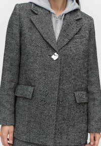 Villa herringbone villakangastakki mustavalkoisena, jossa on kärkikaulus, kaksi etutaskua ja kiiltävä nappikoriste. Kerroksittain harmaan hupparin päällä.