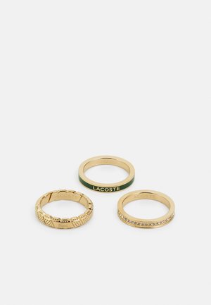 Trois bagues en or : une avec un design texturé, une autre incrustée de vert et gravée "LACOSTE," et la dernière ornée de petites pierres claires.