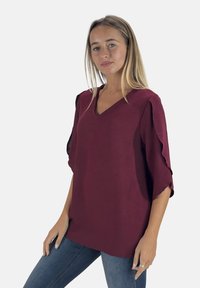 Top color bordeaux con scollo a V, caratterizzato da maniche a volant e tessuto testurizzato. Abbinato a jeans blu, su uno sfondo semplice.