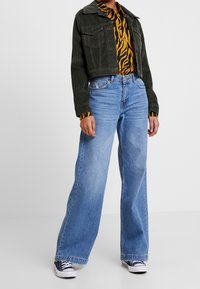 Krótkie czarne kurtka z koralikami nałożona na pasiastą żółto-czarną koszulę, w połączeniu z szerokimi niebieskimi dżinsami i ciemnymi sneakersami.