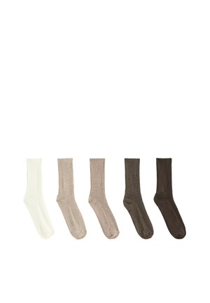 Paire de pieds portant des chaussettes côtelées beige mi-mollet, debout sur une surface blanche sur un fond uni.