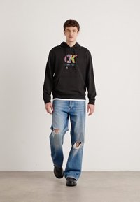 Fekete kapucnis pulóver színes Calvin Klein logóval, világoskék szakadt farmernadrággal és fekete cipővel párosítva. Simább anyag, laza fazon.