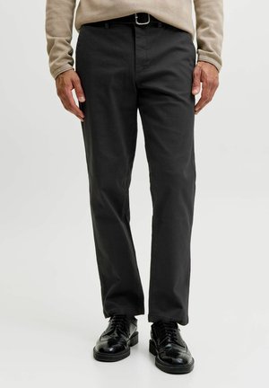 Homme portant un pantalon noir droit avec une ceinture noire, une chemise beige à manches longues et des chaussures habillées noires brillantes, debout sur un fond blanc.