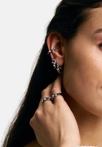 Sudraba rotaslietas ar ģeometriskām formām, tostarp auskariem un gredzeniem ar asiem akcentiem, demonstrētas uz modeļa auss un rokas.