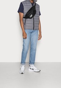 Šedá péřová vesta přes modré tričko, světle modré džíny a bílé a šedé sportovní sneakers. Černá crossbody taška s tištěným logem.