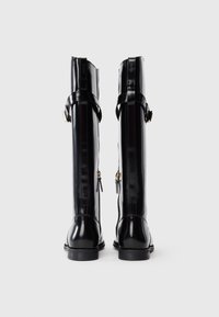 Tall black patent leather boots with side zippers and buckle straps viewed from the back on a white background.