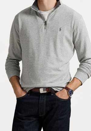 Mężczyzna ubrany w jasnoszary sweter z zamkiem do ćwiartki i logo, ciemne dżinsy, brązowy pasek oraz zegarek na nadgarstku z niebieską tarczą, ręce w kieszeniach.