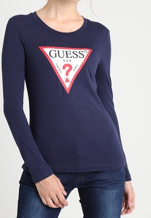 Femme portant un chemisier bleu marine à manches longues avec un grand logo triangulaire Guess rouge et blanc sur la poitrine, et un jean bleu foncé.