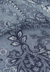 Blå mönstrad textil med intrikata blommönster i olika nyanser av blått och vitt, med en texturerad yta och fransade kanter.