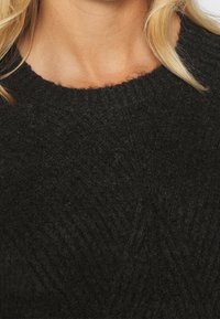 Pull noir tricoté avec une texture côtelée, présentant un col rond et un motif chevrons subtil sur le tissu.