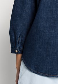 Camicia in denim scuro con una texture liscia, dotata di un orlo arrotondato, maniche arrotolate e cuciture visibili di un colore contrastante.