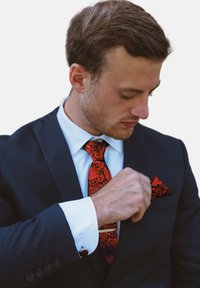 Traje negro con una camisa blanca, con una corbata de paisley rojo vibrante y un pañuelo de bolsillo a juego. Gemelos visibles en el puño de la camisa.