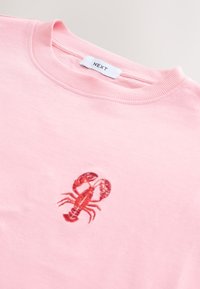 Lyserød sweatshirt har rund halsudskæring og et fremtrædende rødt broderet hummelmotiv på forsiden, med ribstrikket tekstur ved halsudskæringen.