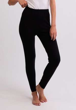 Kvinde iført tætsiddende sorte leggings og en hvid top, stående barfodet mod en ensfarvet lys baggrund.
