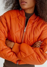 Veste bomber matelassée de couleur orange vif, dotée d'une fermeture éclair argentée et de poignets côtelés. Le matériau semble lisse et léger.