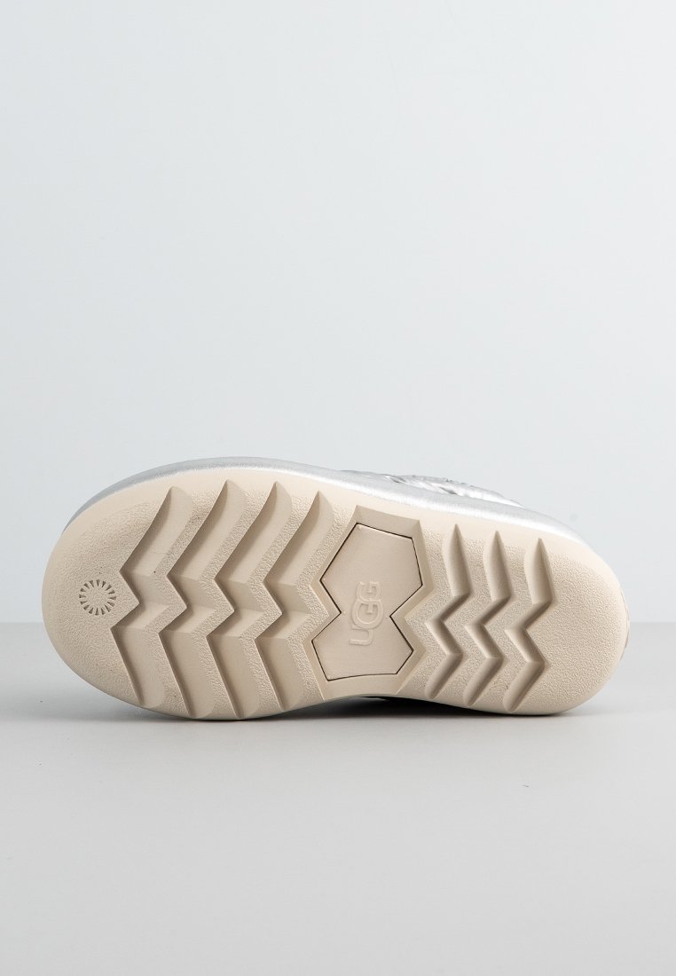 Suola di una scarpa UGG beige con profonde scanalature a zigzag e logo esagonale al centro, appoggiata su una superficie grigia.