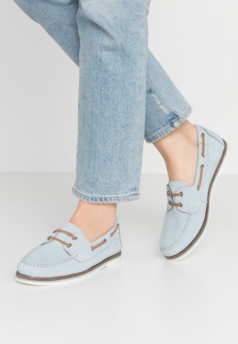 Mocassins en cuir bleu clair avec lacets marron portés avec un jean en denim bleu clair sur un sol blanc.