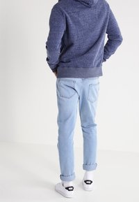 Person mit blauem Kapuzenpullover, hellblauen hochgekrempelten Jeans, weißen Socken und weißen Nike-Sneakern, steht vor einem schlichten hellen Hintergrund.