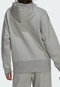 Personne portant un sweat à capuche gris clair et un pantalon de survêtement assorti, vue de dos avec les mains reposant le long du corps.