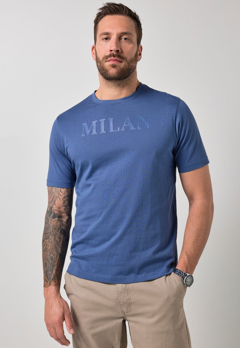 Homme aux cheveux courts et barbe, portant un t-shirt bleu "MILAN" et un pantalon beige, debout avec une main dans la poche et un tatouage visible sur le bras.