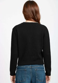 Donna con lunghi capelli castani, che indossa un maglione nero e jeans blu, in piedi di spalle contro uno sfondo bianco uniforme.