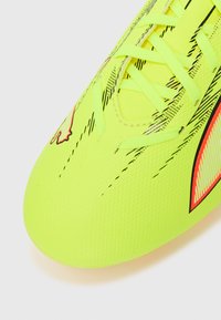 Żółty but piłkarski z teksturowaną powierzchnią, ozdobiony cienkimi czarnymi liniami i pomarańczowymi akcentami wzdłuż boków. Sznurowadła są jaskrawo żółte.