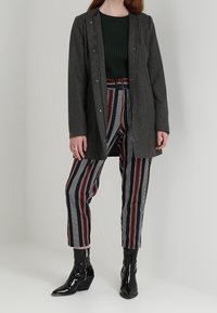Abrigo gris oscuro con cierres de broches, suéter de punto verde texturizado, pantalones a rayas en rojo, negro y blanco, combinados con botas negras de charol hasta el tobillo.
