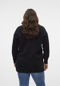 Osoba z długimi falującymi brązowymi włosami, ubrana w czarny sweter i niebieskie dżinsy, stojąca tyłem na jednolitym białym tle.