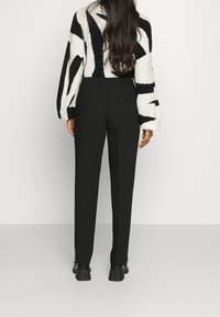 Nainen, jolla on pitkät tummat hiukset, pukeutunut mustiin mittatilauspö trousers, mustiin kenkiin ja valkoiseen neuleeseen, jossa on rohkeita mustia abstrakteja raitoja, seisoo valkoista seinää vasten.