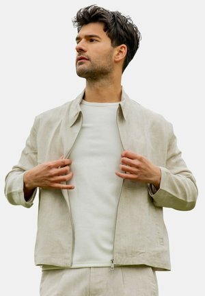 Homme aux cheveux foncés portant une veste beige clair sur une chemise blanche, tenant la veste ouverte, regardant vers sa gauche sur un fond uni.