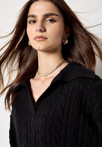 Suéter negro de punto trenzado con cuello, presenta un patrón texturizado. Un collar dorado y pendientes de aro complementan el conjunto. Cabello largo y ondulado.