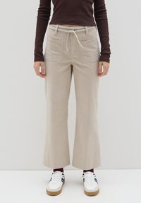 Beige wijde broek gemaakt van zachte stof, met een trekkoord in de taille en twee voorkuilen. Gepaard met witte sneakers en bordeauxrode sokken.