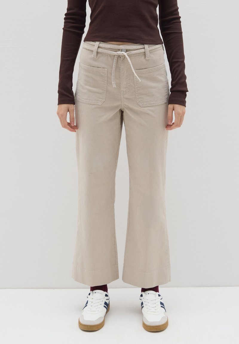 Beige wijde broek gemaakt van zachte stof, met een trekkoord in de taille en twee voorkuilen. Gepaard met witte sneakers en bordeauxrode sokken.
