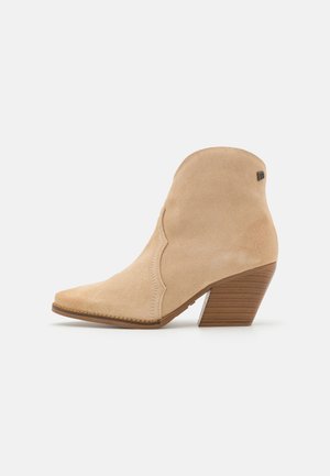 Botín de ante beige con punta afilada, diseño ondulado y tacón de madera apilada. Presenta un sutura sutil y un pequeño detalle de logo.