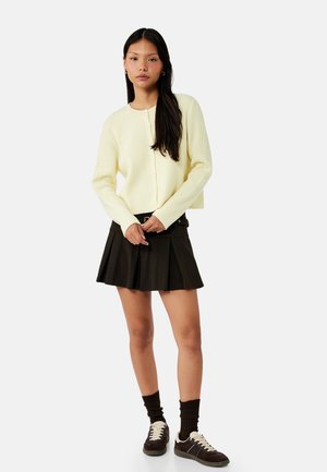 Joven mujer de pie con un cárdigan abotonado amarillo pálido, falda negra plisada, calcetines negros y zapatillas negras.