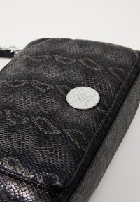 Črn usnjen crossbody nahrbtnik z srebrno okroglo oznako in subtilnim vzorčnim dizajnom na enotni podlagi.