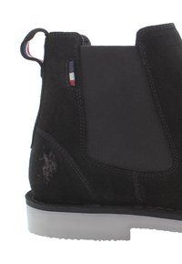 Botín Chelsea de ante negro con paneles laterales elásticos, una lengüeta y suela de goma blanca contrastante. Presenta un logotipo grabado en el talón.