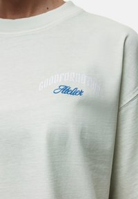 Felpa verde chiaro realizzata in morbido tessuto, con ricamo bianco e blu della scritta "GOODFORNOTHING" e "Atelier" sul petto.