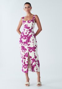 Robe maxi à imprimé floral crème et violet avec décolleté carré, corsage ajusté, boutons sur le devant et fente latérale. Portée avec des sandales tressées.