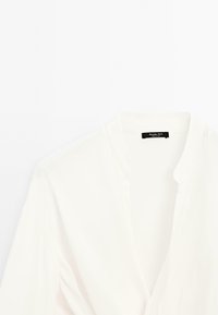Blouse blanche avec un décolleté en V, une texture douce et un design minimaliste. Présente un léger éclat et une étiquette avec le nom de la marque au niveau du col.