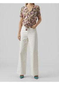 Donna che indossa una blusa a maniche corte con bottoni a fiori annodata in vita, pantaloni bianchi a gamba larga e scarpe verde acqua a punta con fibbie.