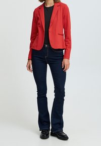 Giacca rossa con un solo bottone, rever a contrasto e tasche laterali, abbinata a jeans svasati blu scuro e scarpe nere.