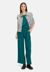 Tweed jasje met zwarte knopen over een smaragdgroene jumpsuit met een gestrikte taille. Model draagt zwarte enkellaarsjes en staat tegen een witte achtergrond.