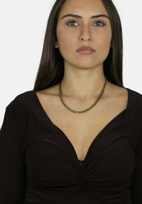 Złoty łańcuszek na modelce noszącej brązową bluzkę z głębokim dekoltem w kształcie V i węzłem na środku, o gładkiej fakturze.