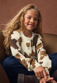 Pleteni kardigan v kremni barvi z rjavim kravjim potiskom, z okroglim izrezom in zapiranjem na gumbe. Nošen z temi modrimi kavbojkami in rjavimi škornji.