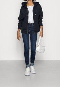 Felpa con zip color navy e cappuccio, abbinata a una t-shirt bianca, jeans scuri, sneakers bianche e una borsa a tracolla bianca trapuntata.